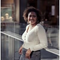 Ane Ogbe. Credit: Ane Ogbe Photo of Ane Ogbe standing against a glass screen. Ane is wearing a white cardigan and holding her glasses.