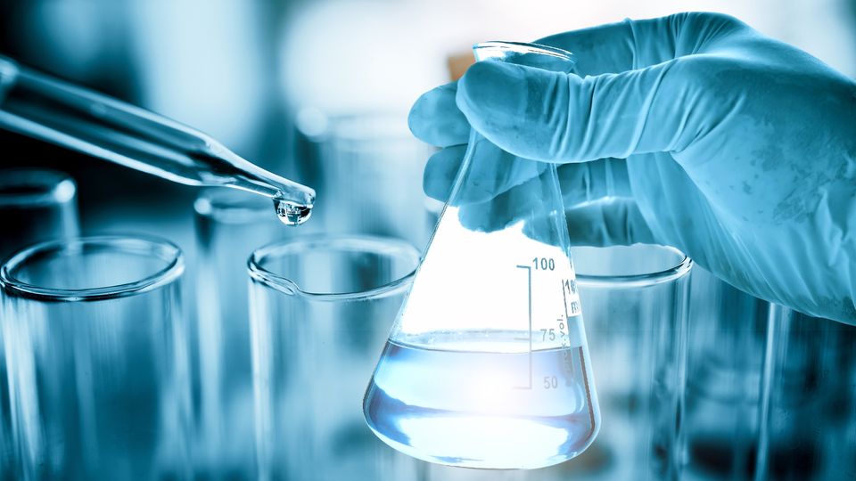 Gloved hand holding a conical flask with blue solution near test tubes, illustrating molarity vs molality.