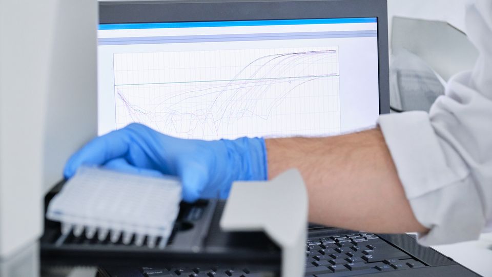 A researcher takes a 96-well plate out of a machine. A screen with a graph is in the background.