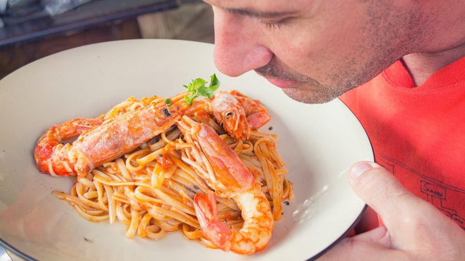Man in red shirt smells a dish containing seafood pasta.