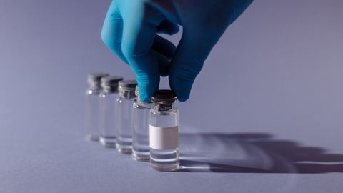 A gloved hand holding a clear vial filled with liquid with a white label between two fingers. Four clear vials sit behind.