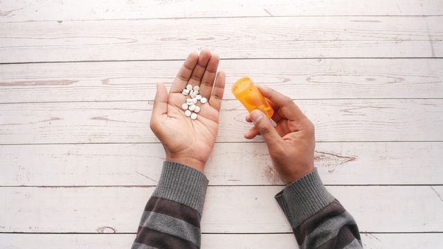 Two arms extended on a white wooden surface. The right hand is holding an empty medication tube. Many white tablets can be seen on the palm of the left hand. 