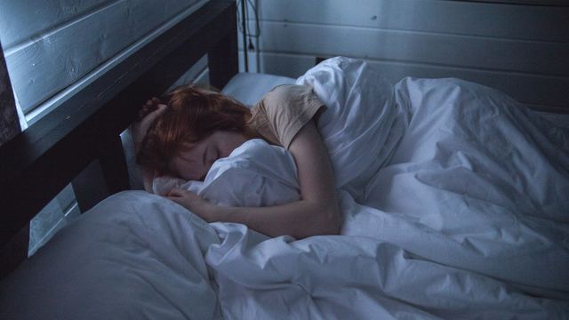 A person sleeping in a dark room in a bed with a dark wooden frame and a white duvet. 