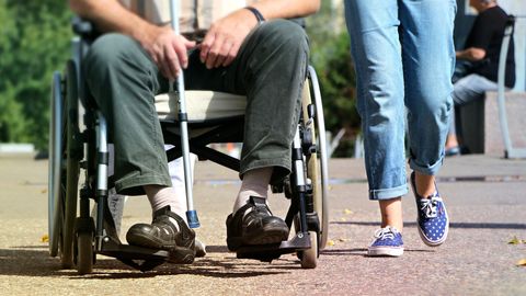 A young person wearing blue jeans and sandshoes walks next to an older person in a wheelchair.