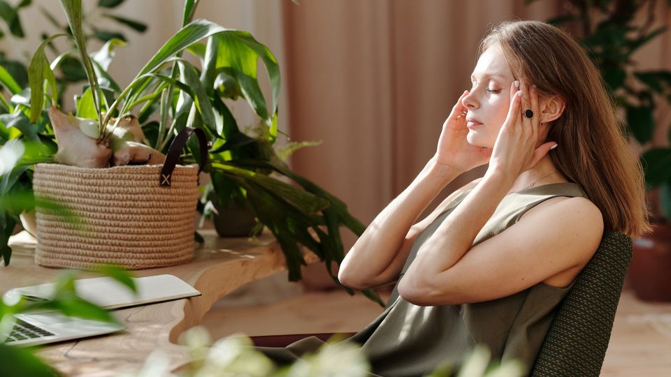 A woman sits at a desk with her eyes closed and fingers rubbing her temples. 