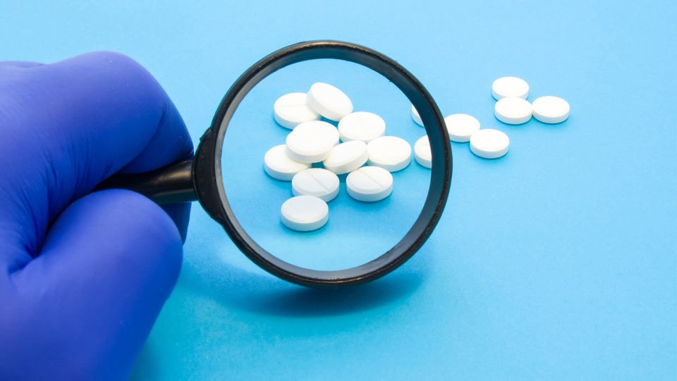 A gloved hand holds a small magnifying glass over white, disc-shaped pills. 