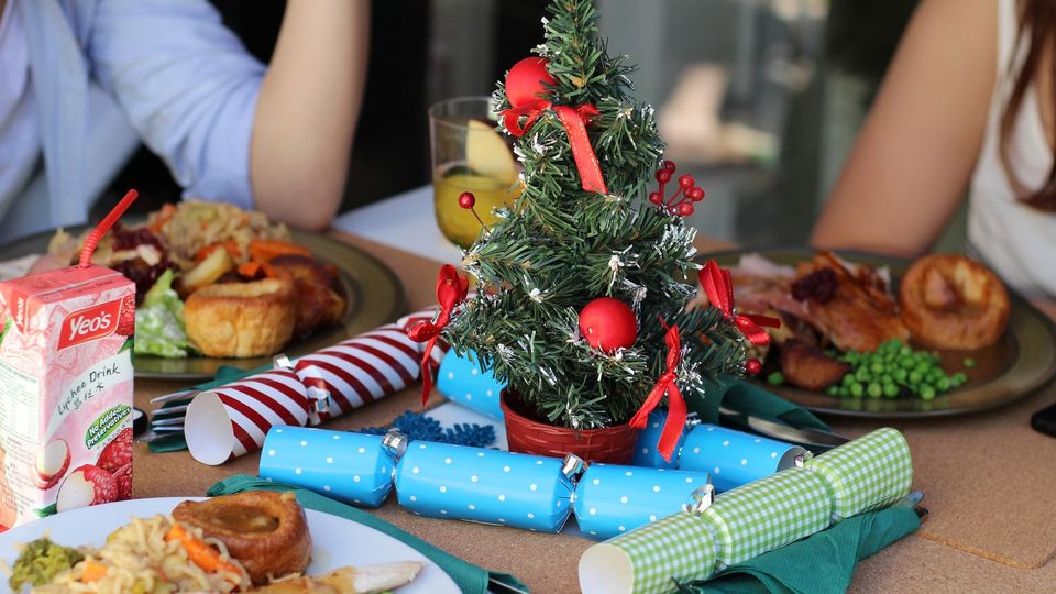 People sit at a table eating Christmas dinner and a small Christmas tree and crackers on the table.