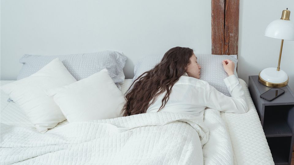 A woman with long brown hair lies asleep on her front in a bed with cream sheets. 