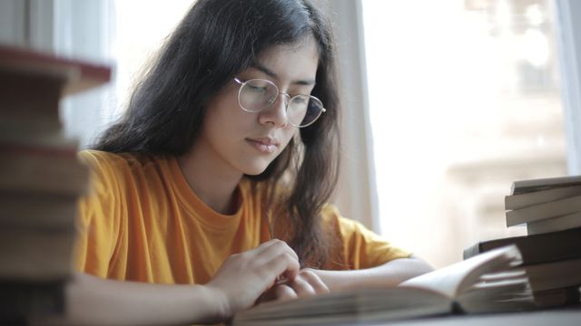 A girl wearing a yellow t-shirt and round glasses sits at a table and reads a book.