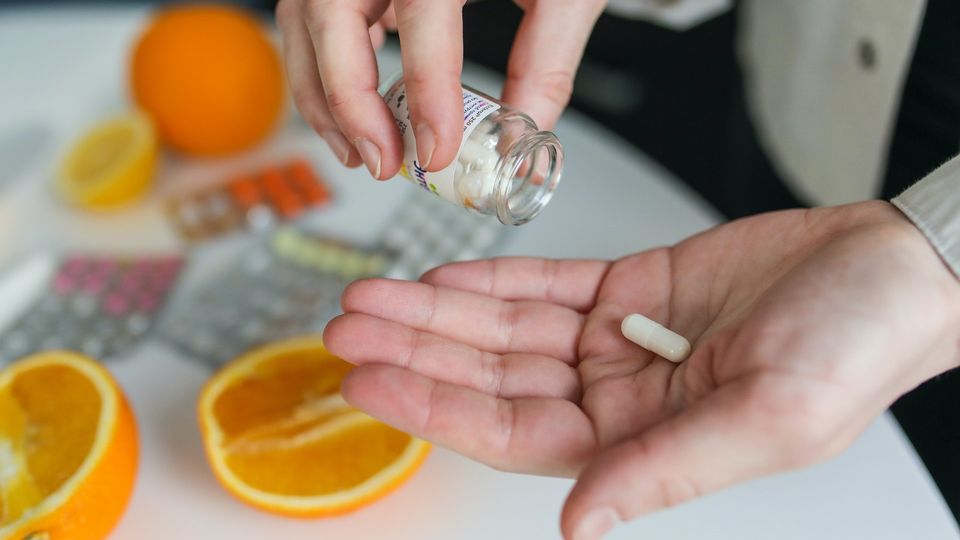 A closeup of a tablet in the palm of hand, shaken from a bottle in the other. Oranges and other packs of tablets can be seen on a table in the background.