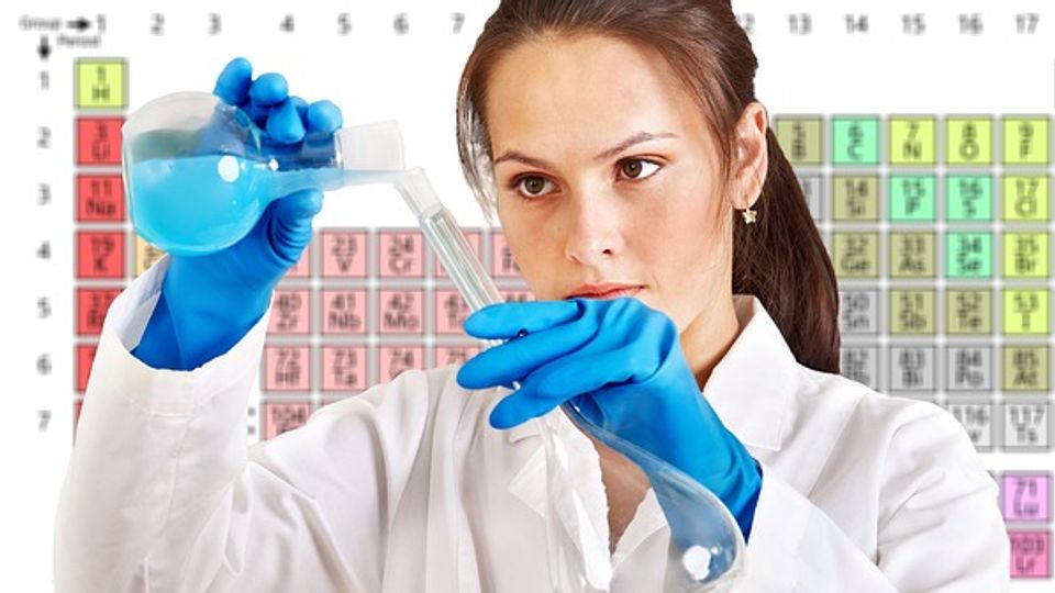 Woman in a lab coat stood in front of the periodic table pouring liquid between two glass flasks.