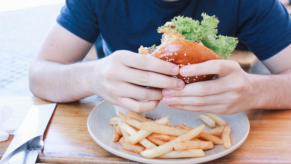 A man in a blue t-shirt holds a burger above a plate of chips.