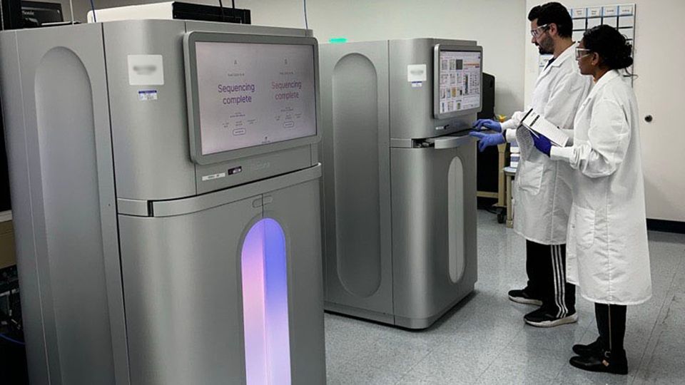 Two scientists in lab coats standing in front of sequencing machines in a lab.