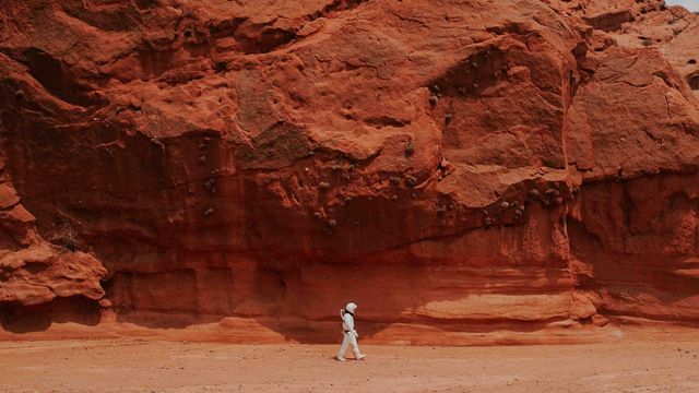 Person in spacesuit near red rocks.