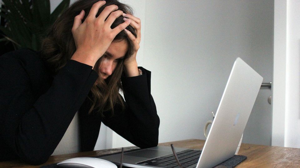 A woman sits at a laptop, hands on her head.