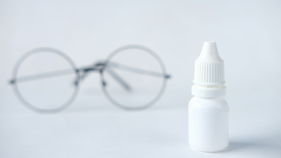 A bottle of eye drops with glasses in the background.