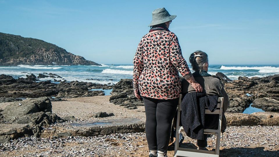 A pair of women on the beach, one sitting on a wooden chair and the other standing.