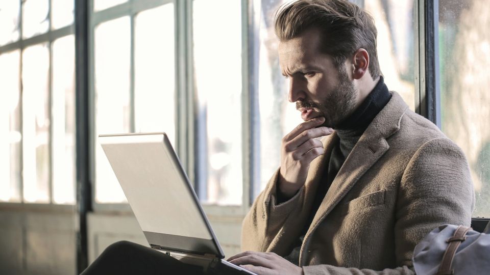 A man in a tan jacket and black turtleneck sits thinking and working on a laptop.