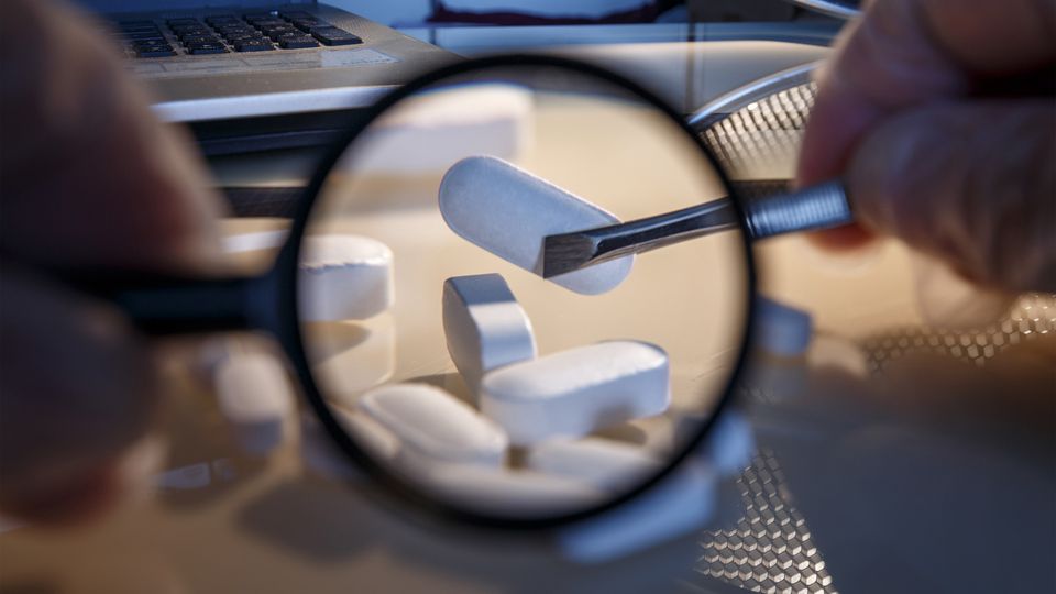 A white tablet held with tweezers seen through a magnifying glass