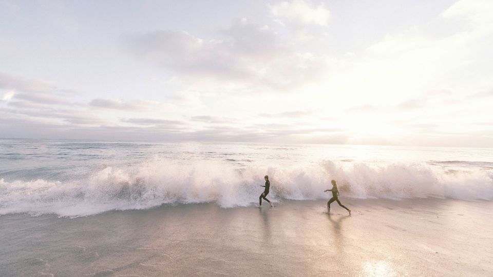 Two people in wetsuits run towards the sea.