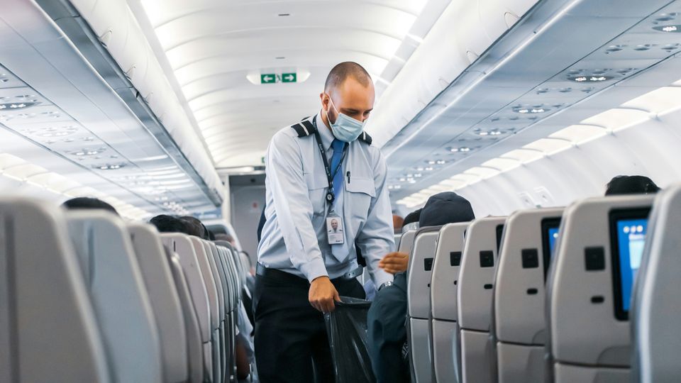 An airport worker on a plane wearing a mask collecting rubbish from a passenger