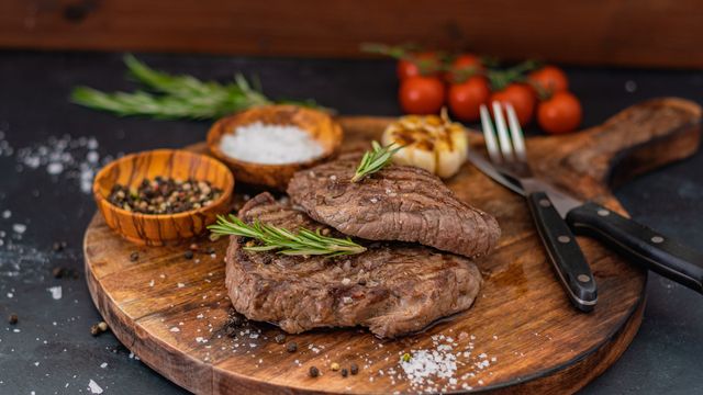 Steak on a wooden board with two small bowls of salt and pepper.