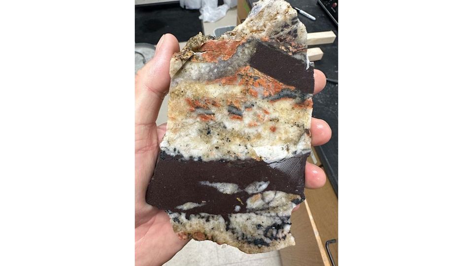 A hand holds a large rock with bands of different colors in it.
