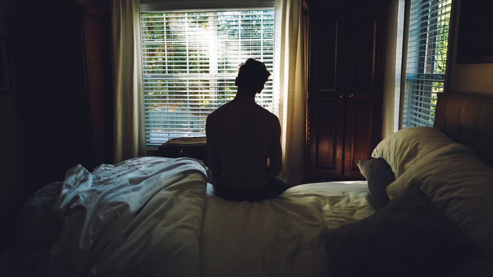 A man sitting on the edge of a bed looking out the window.