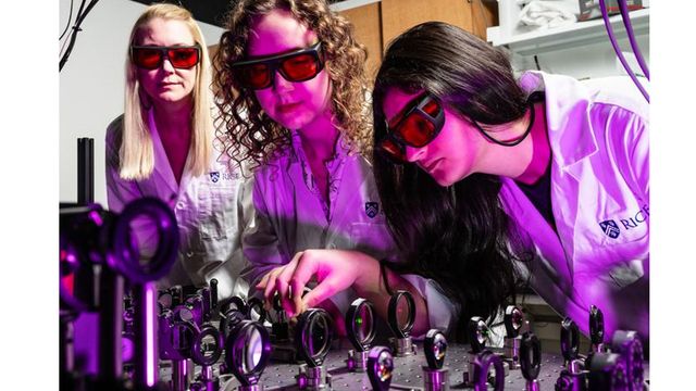 Three researchers wearing red-tinted goggles look at a series of microscope lenses.