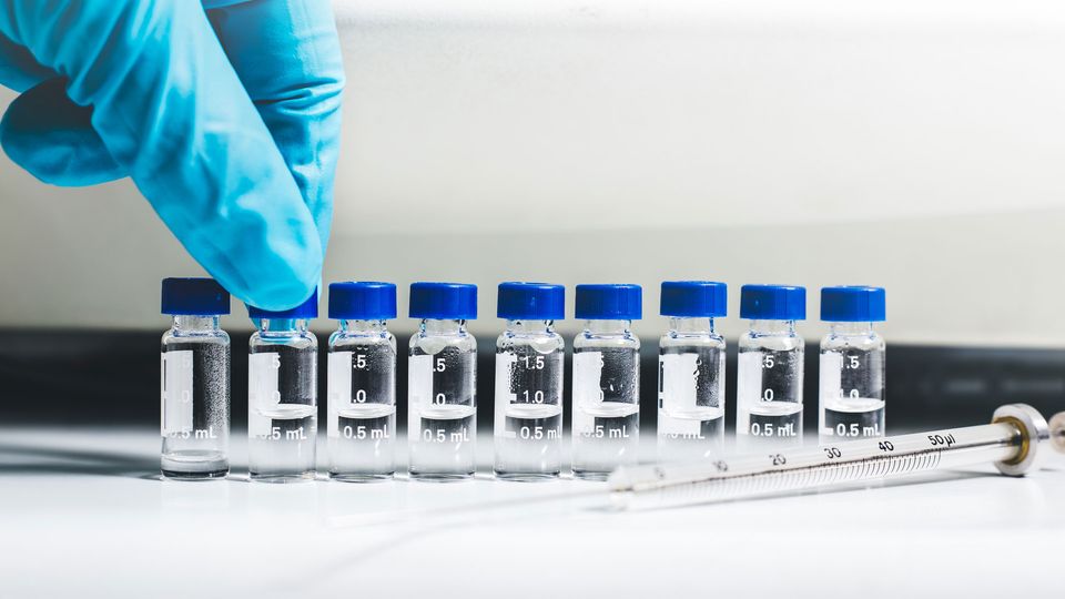 Row of sample vials with blue caps used in mass spectrometry, next to a glass syringe on a laboratory surface.