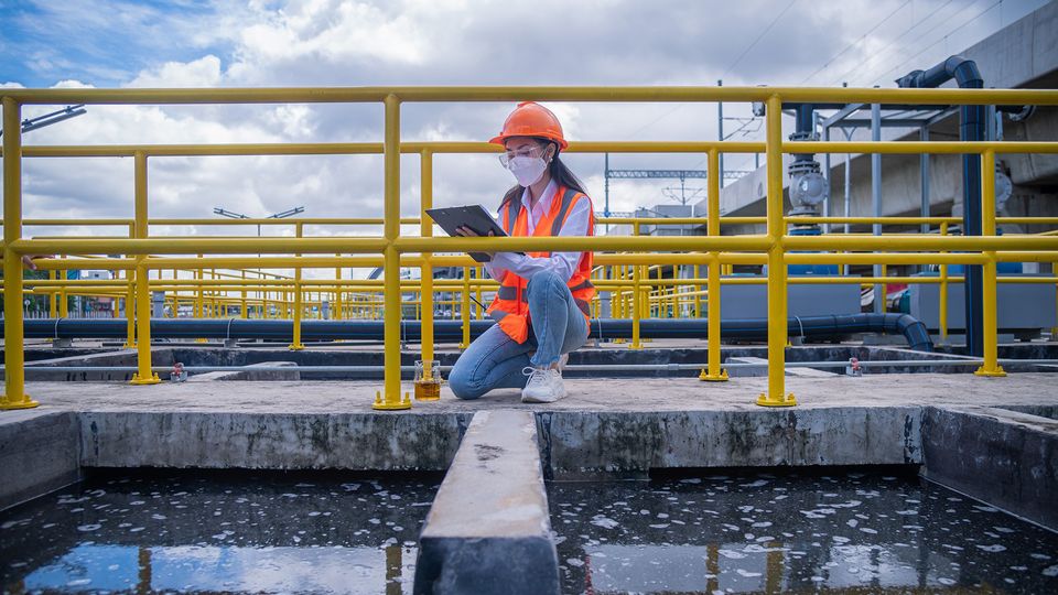 A researcher in a hard hat and safety vest conducting environmental monitoring at a wastewater treatment facility