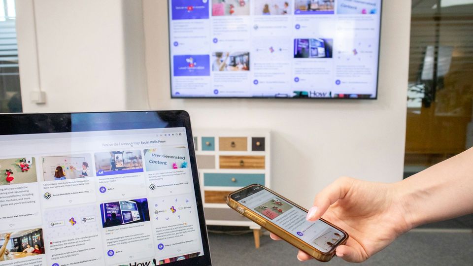 A person holding a phone, with a laptop in front of them and a TV on the wall behind.