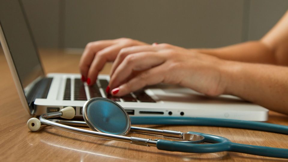 A person types on a laptop, with a stethoscope next to them.