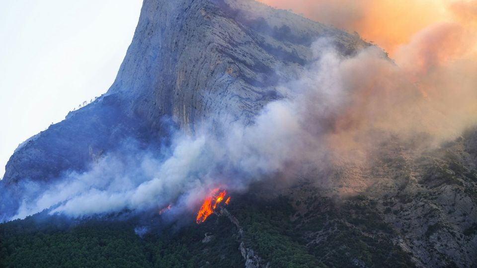 A wildfire breaks out in the woods on a mountain. Light from another fire is visible over the peak.
