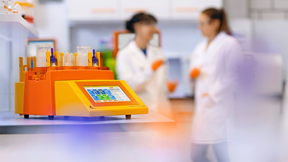 Image of a orange and yellow lab instrument with a digital touchscreen and test tubes on top is prominently shown on a workbench, with two scientists in white lab coats.