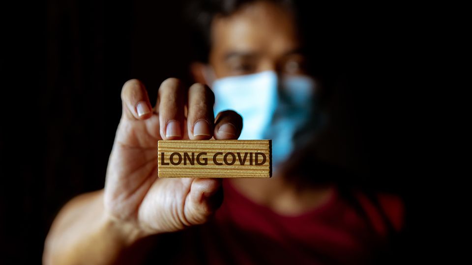 Man in a mask holding up a small wooden sign with "long COVID" written across it.