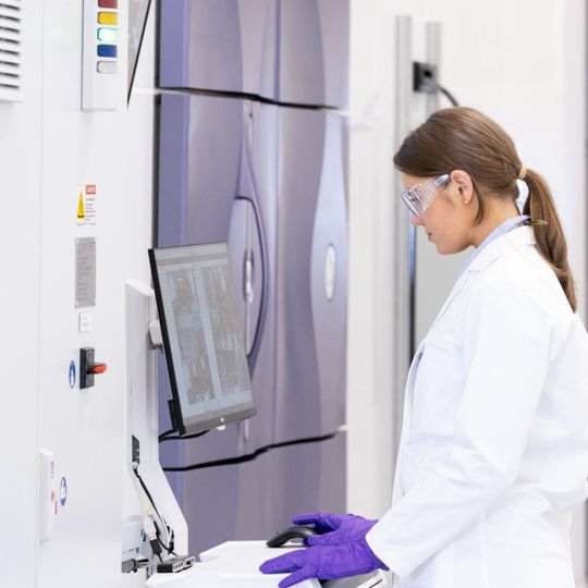 A female scientist in a white lab coat and purple gloves operating an ultra low freezer. 