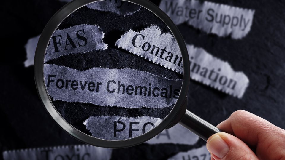 A person holds a magnifying glass over scraps of paper that read "PFAS", "forever chemicals", "contamination" and other keywords.