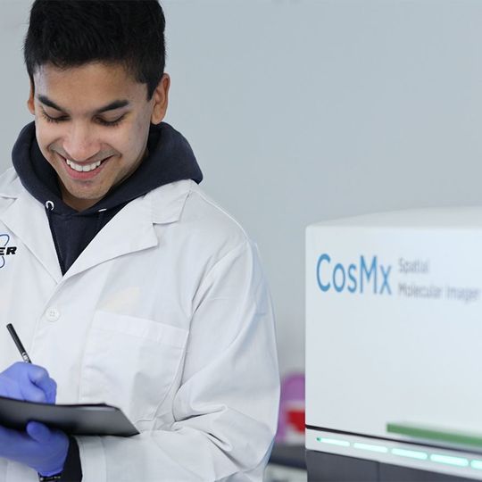 Scientist in a Bruker lab coat smiles while writing on a clipboard next to a CosMx Spatial Molecular Imager 