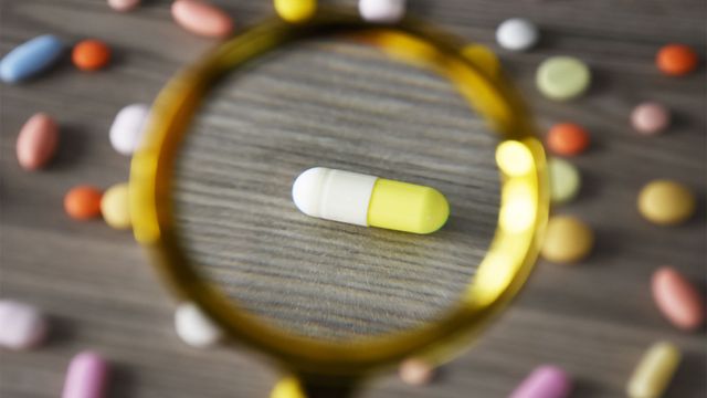 Yellow and white capsule pill focused under a magnifying glass surrounded by various colorful pills on a wooden surface.
