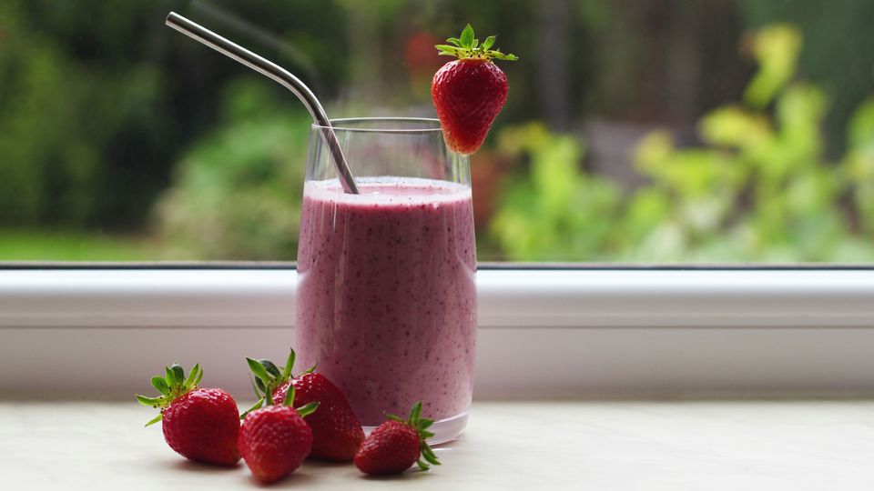 A strawberry smoothie in a glass, near a window, whole strawberries littered nearby.