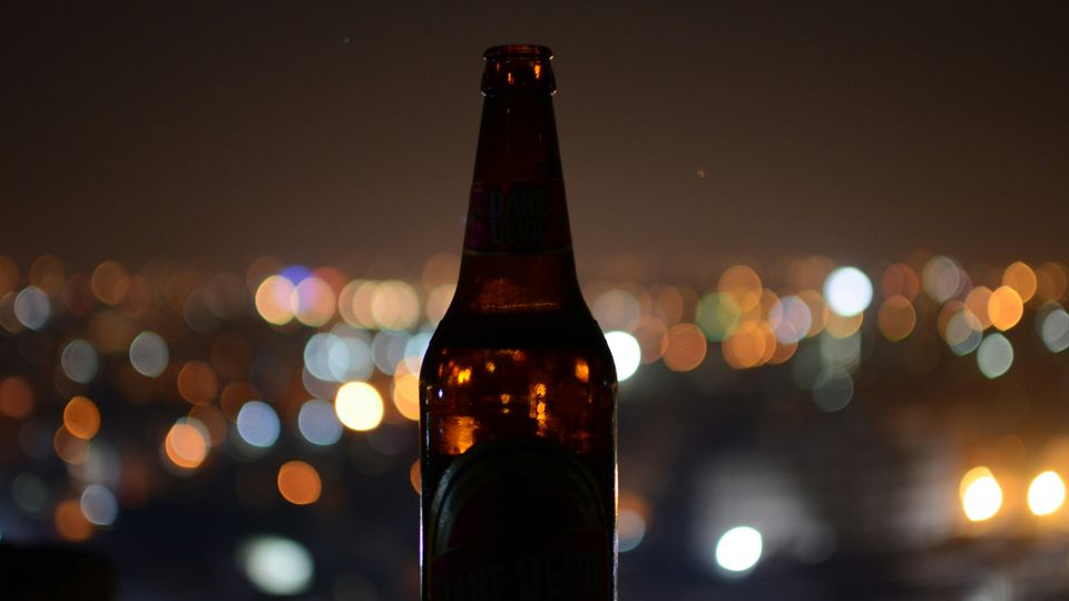 The silhouette of a beer bottle against the blurry lights of a city skyline at night.