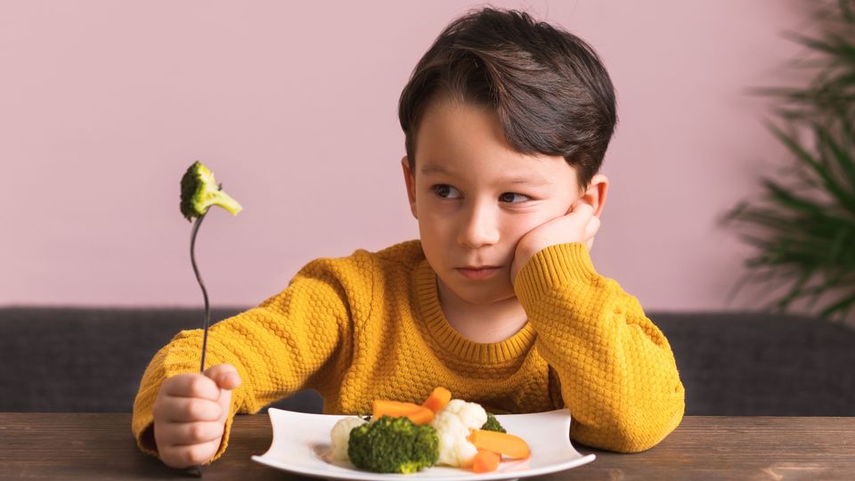 Young boy showing reluctance while holding a fork with broccoli, symbolizing fussy eating habits.