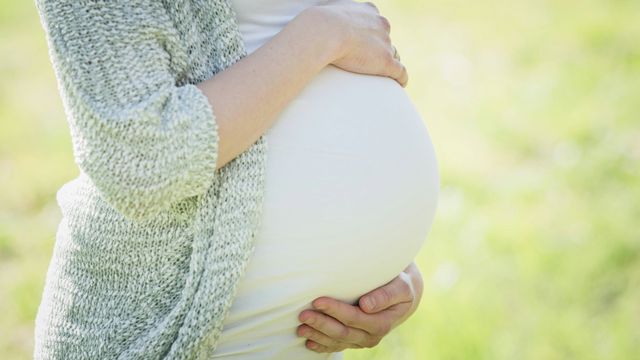 A pregnant woman in a white t-shirt and blue cardigan holding her stomach.