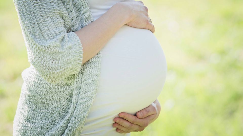 A pregnant woman in a white t-shirt and blue cardigan holding her stomach.
