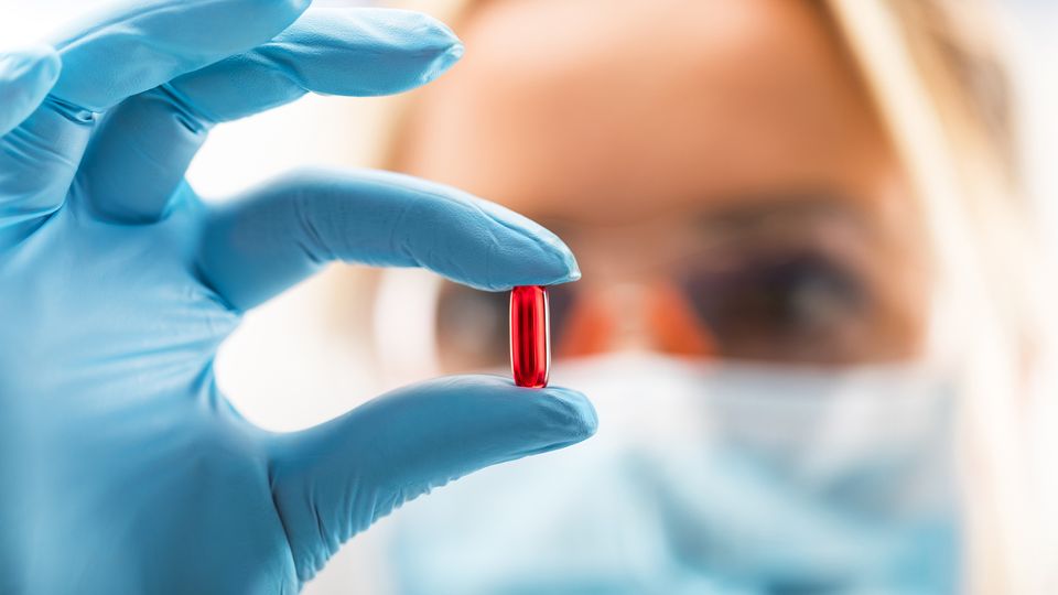 A researcher wearing blue sterile gloves holds up a small red capsule pill.