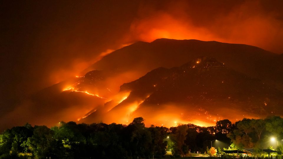 A hill at night, lit by a forest fire. Treeline and electric lights visible in the foreground.