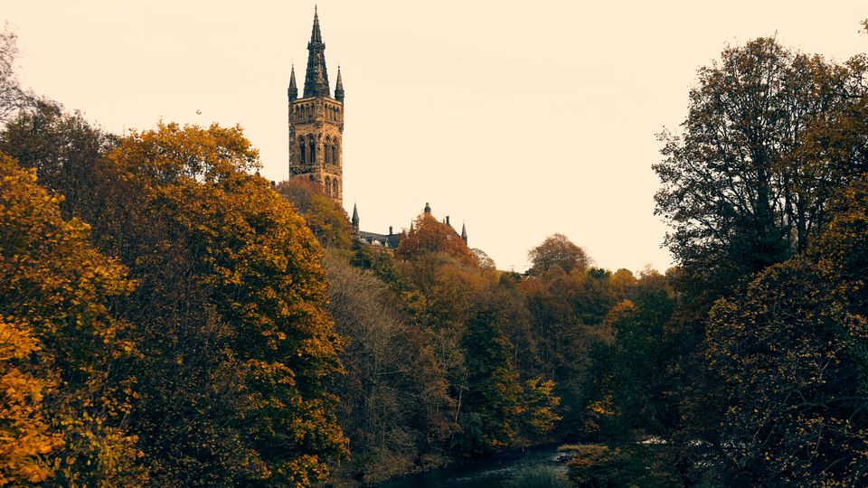 The spire of Glasgow University protruding through the autumnal treeline. River visible. Overcast day.
