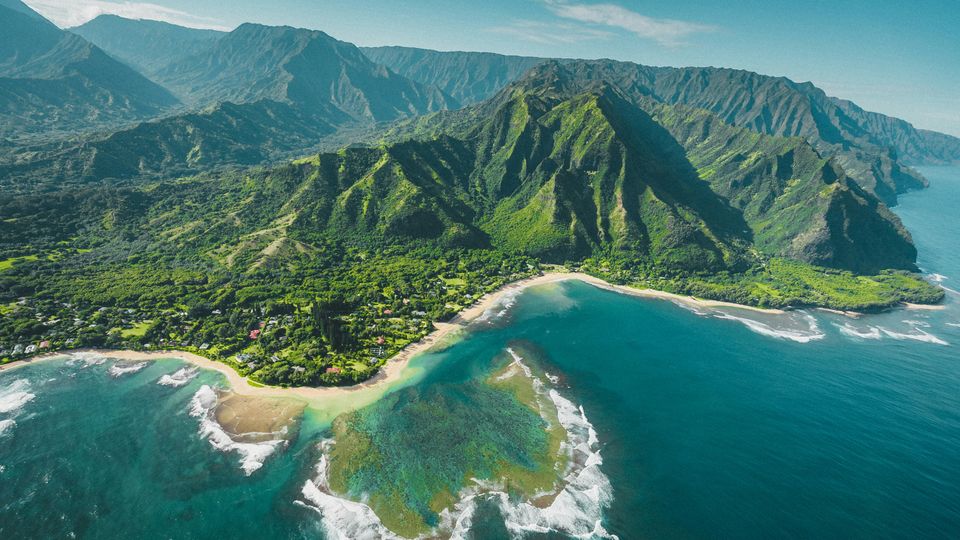 A green, mountainous island from above, coastline and surf can be seen.