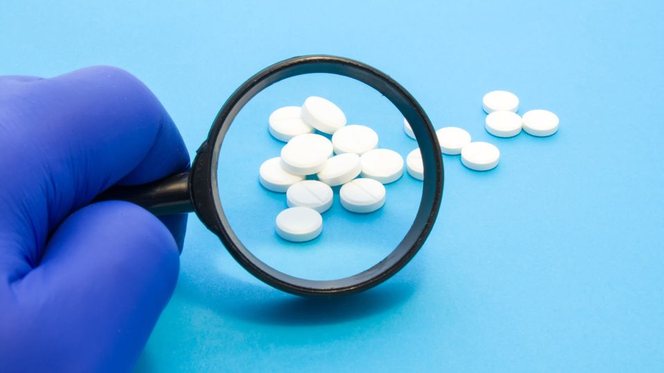 Gloved hand holding magnifying glass over white paracetamol tablets on a blue background.